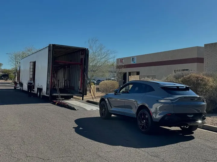 Grey Aston Martin DBX seen from behind, beside an enclosed car transporter outside the shop