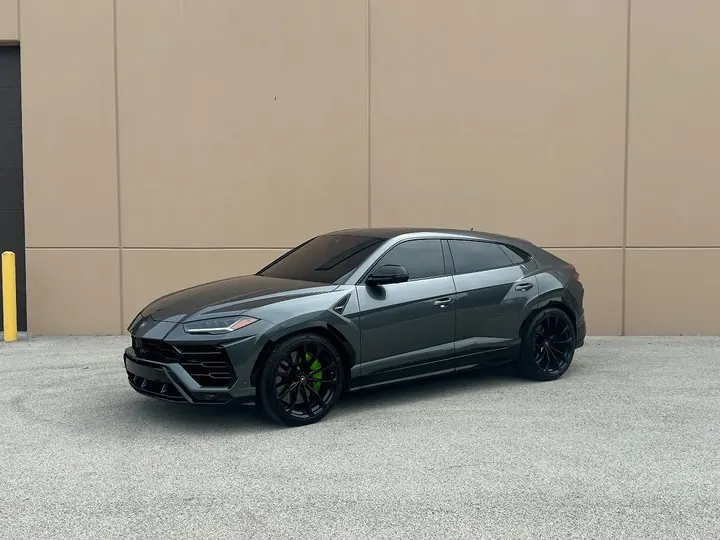 Grey Lamborghini Urus with black wheels and green brake calipers, parked by a tan wall