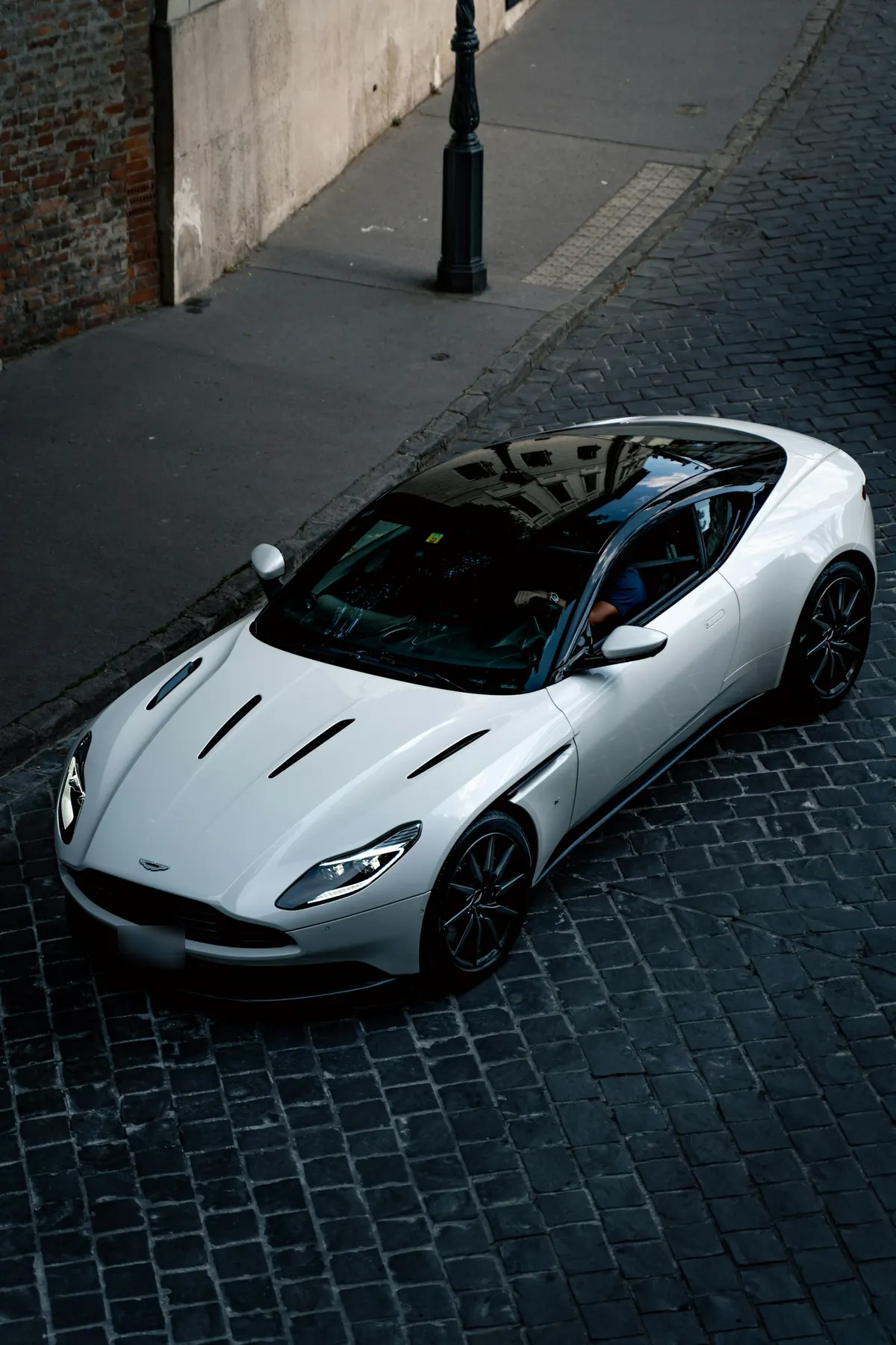 Front three-quarter view from above of a white Aston Martin DB11 V12 on a cobbled street