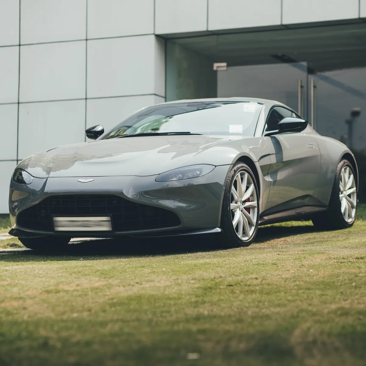 Front three-quarter view of a grey Aston Martin V12 Vantage coupe parked on a lawn outside a building