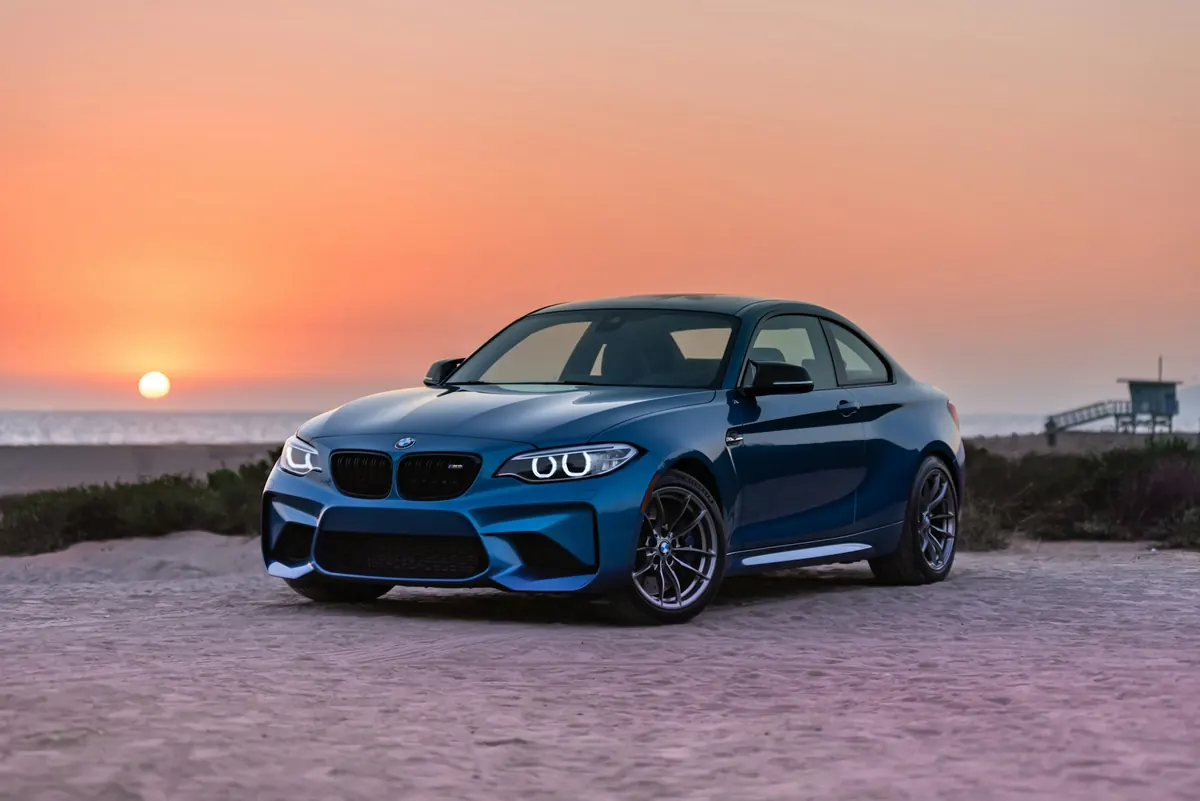Front three-quarter view of a blue BMW M2 (F87) on a beach at sunset