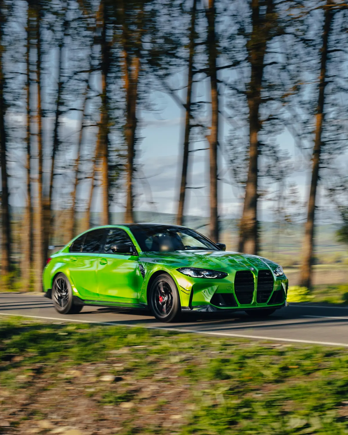 Front three-quarter view of a green BMW M3 Competition (G80) sedan driving on a tree-lined country road