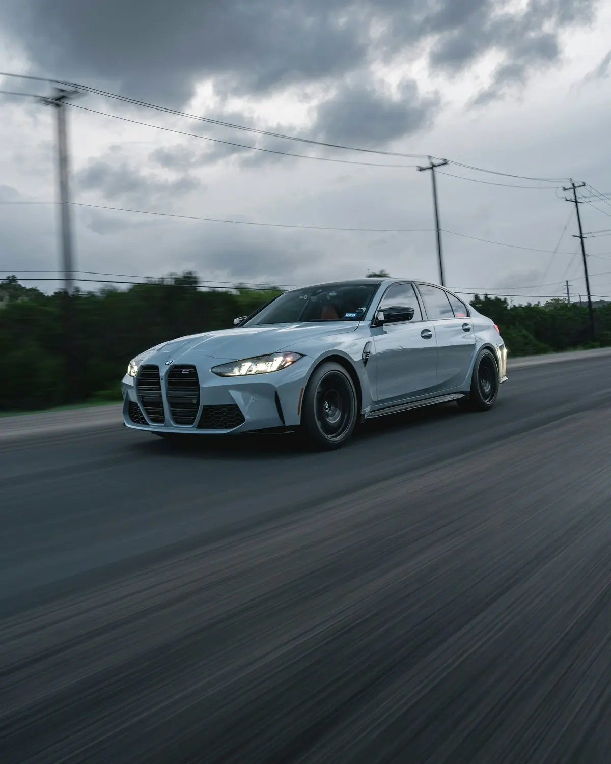 Front three-quarter view of a light grey BMW M3 Competition xDrive (G80) sedan driving on a highway
