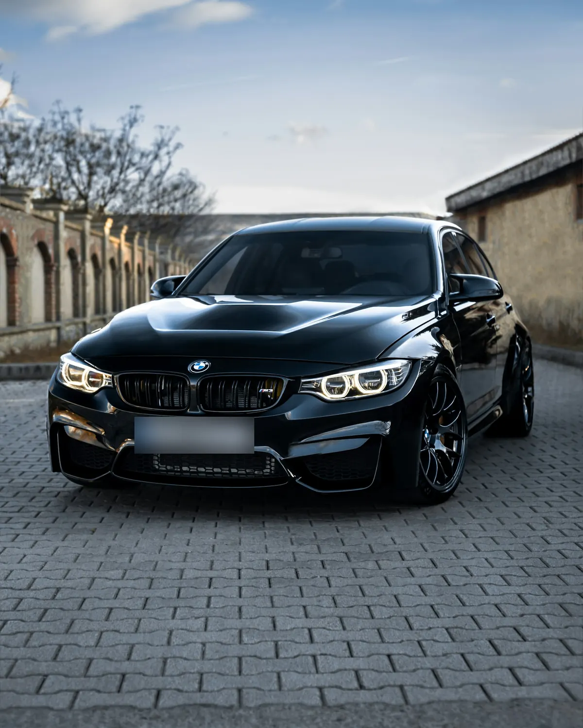 Front three-quarter view of a black BMW M3 (F80) sedan on a cobblestone lane