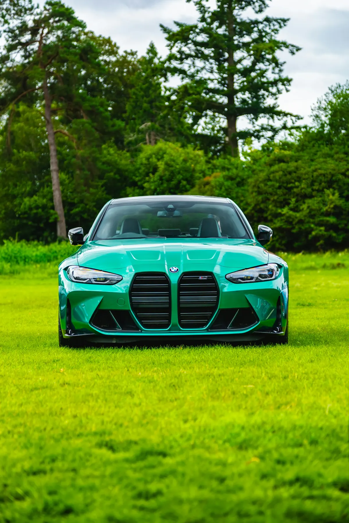Front view of a green BMW M3 (G80) parked on a lawn in front of trees