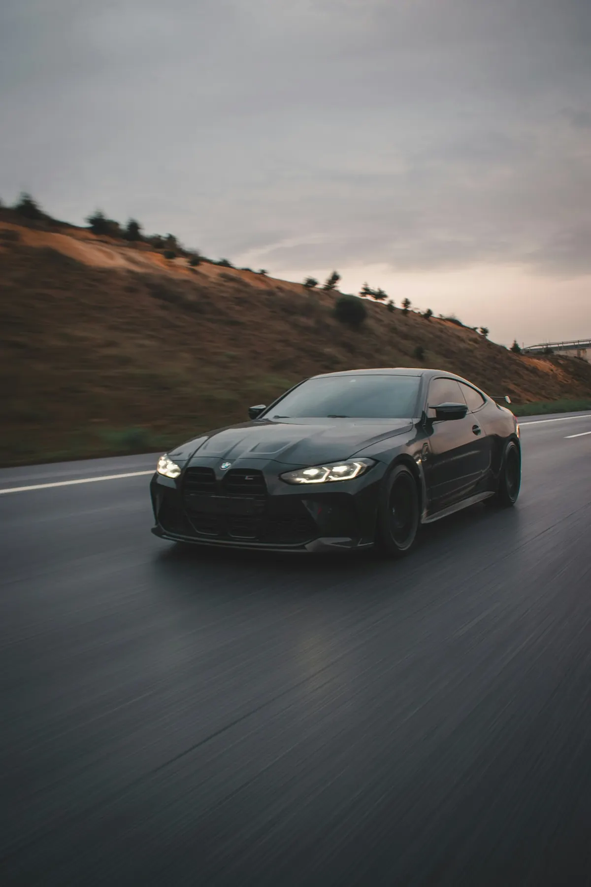 Front three-quarter view of a black BMW M4 Competition (G82) coupe driving on a highway at dusk