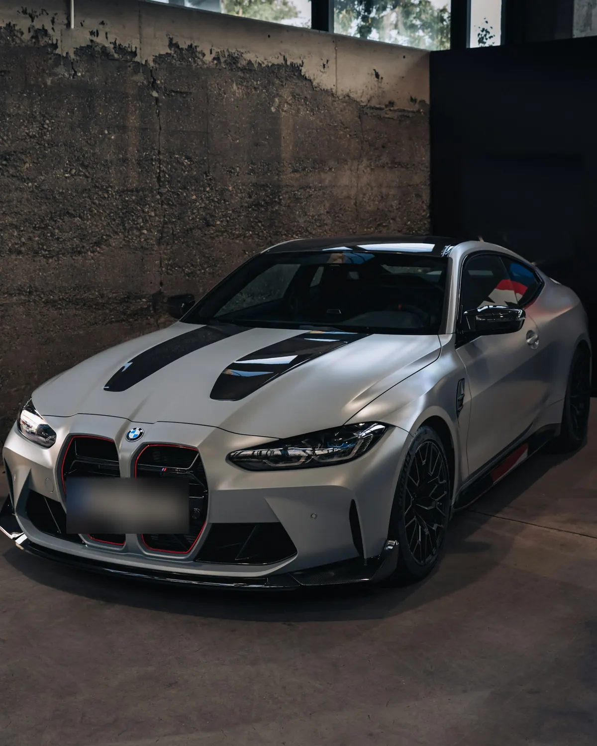 Front three-quarter view of a matte light grey BMW M4 CSL (G82) parked indoors by a concrete wall