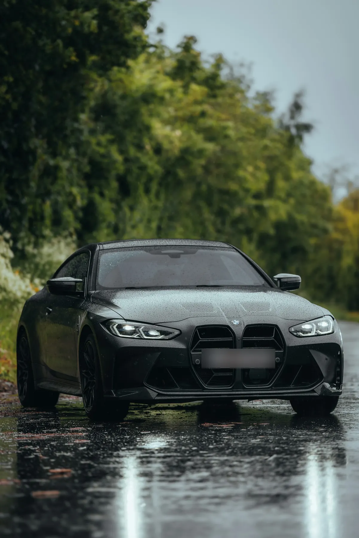 Front three-quarter view of a matte dark grey BMW M4 (G82) coupe on a wet tree-lined road