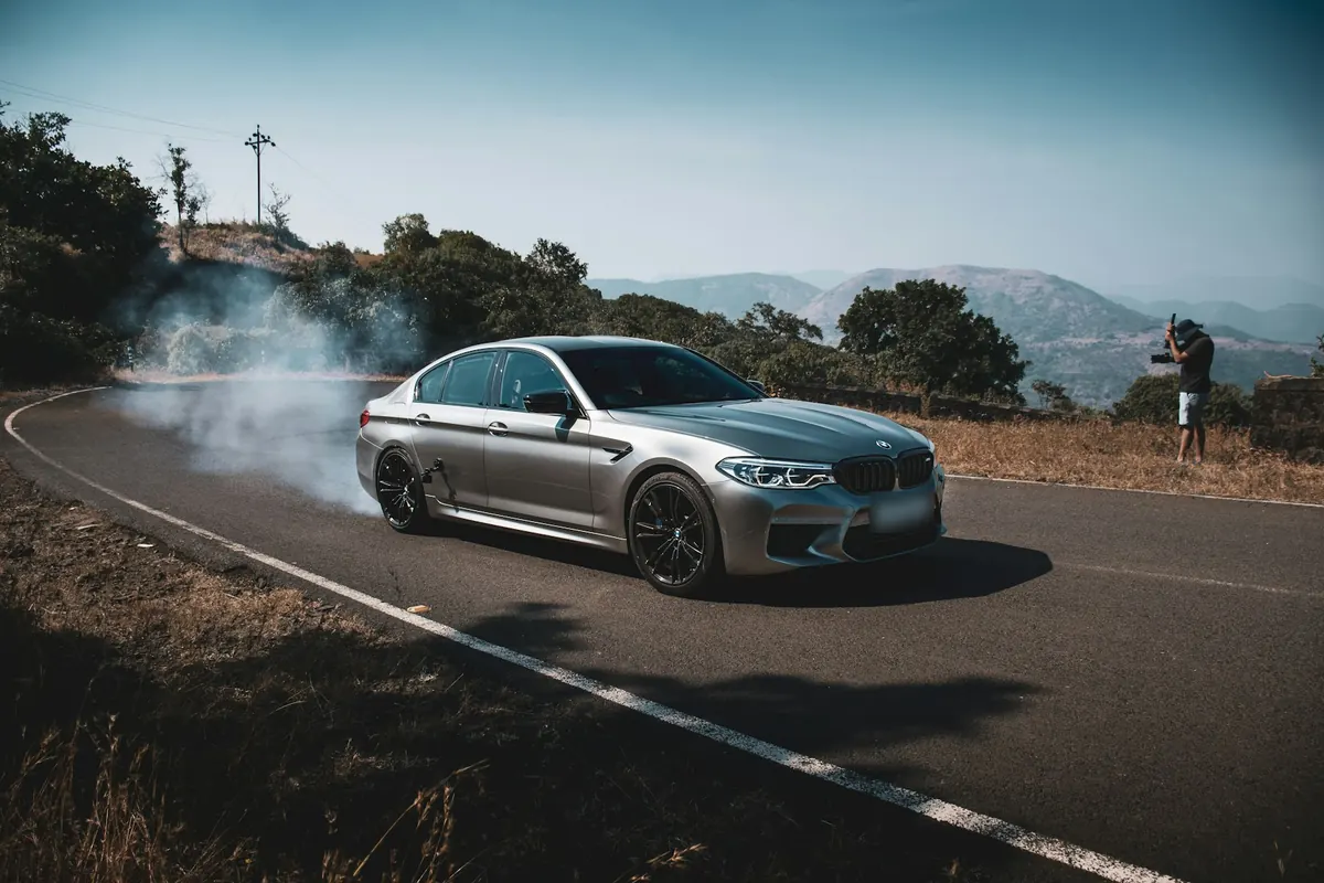 Front three-quarter view of a silver BMW M5 Competition (F90) doing a burnout on a hill road