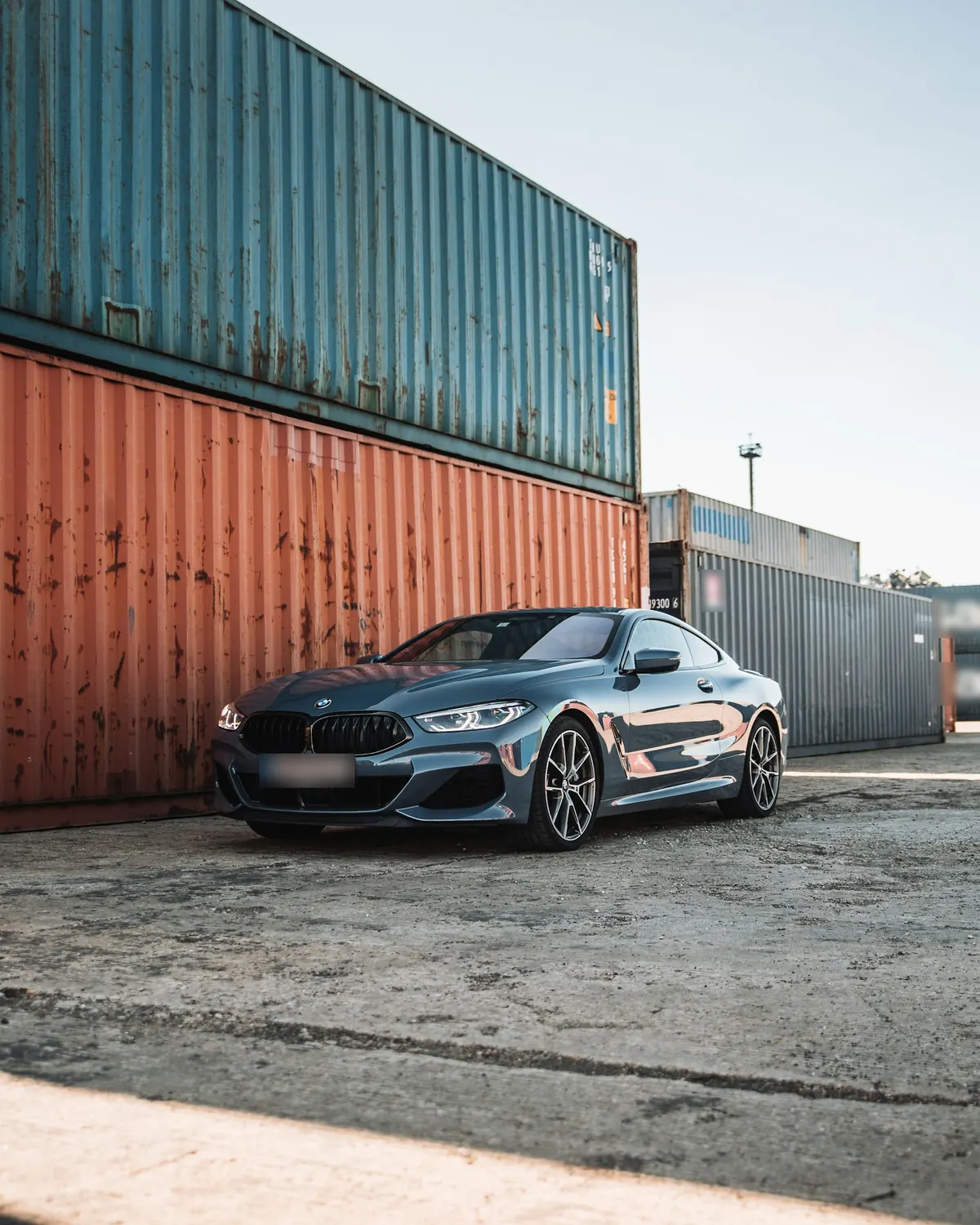 Front three-quarter view of a blue-grey BMW M850i Coupe (G15) between shipping containers