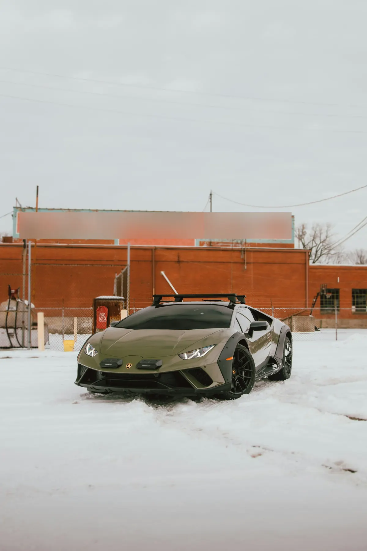 Front three-quarter view of an olive green Lamborghini Huracán Sterrato with a roof rack in the snow