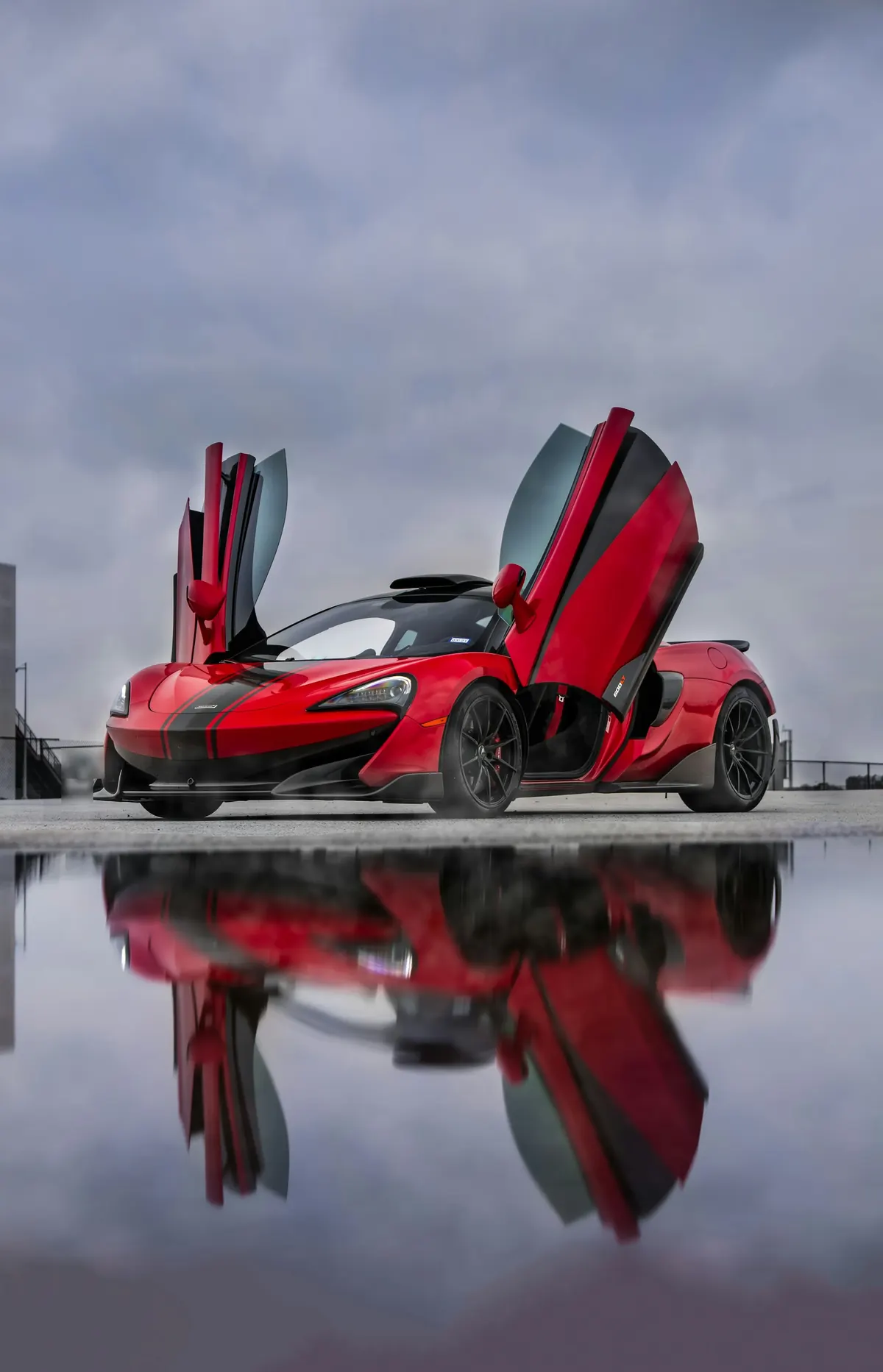 Front three-quarter view of a red McLaren 600LT with its dihedral doors open, reflected in a puddle