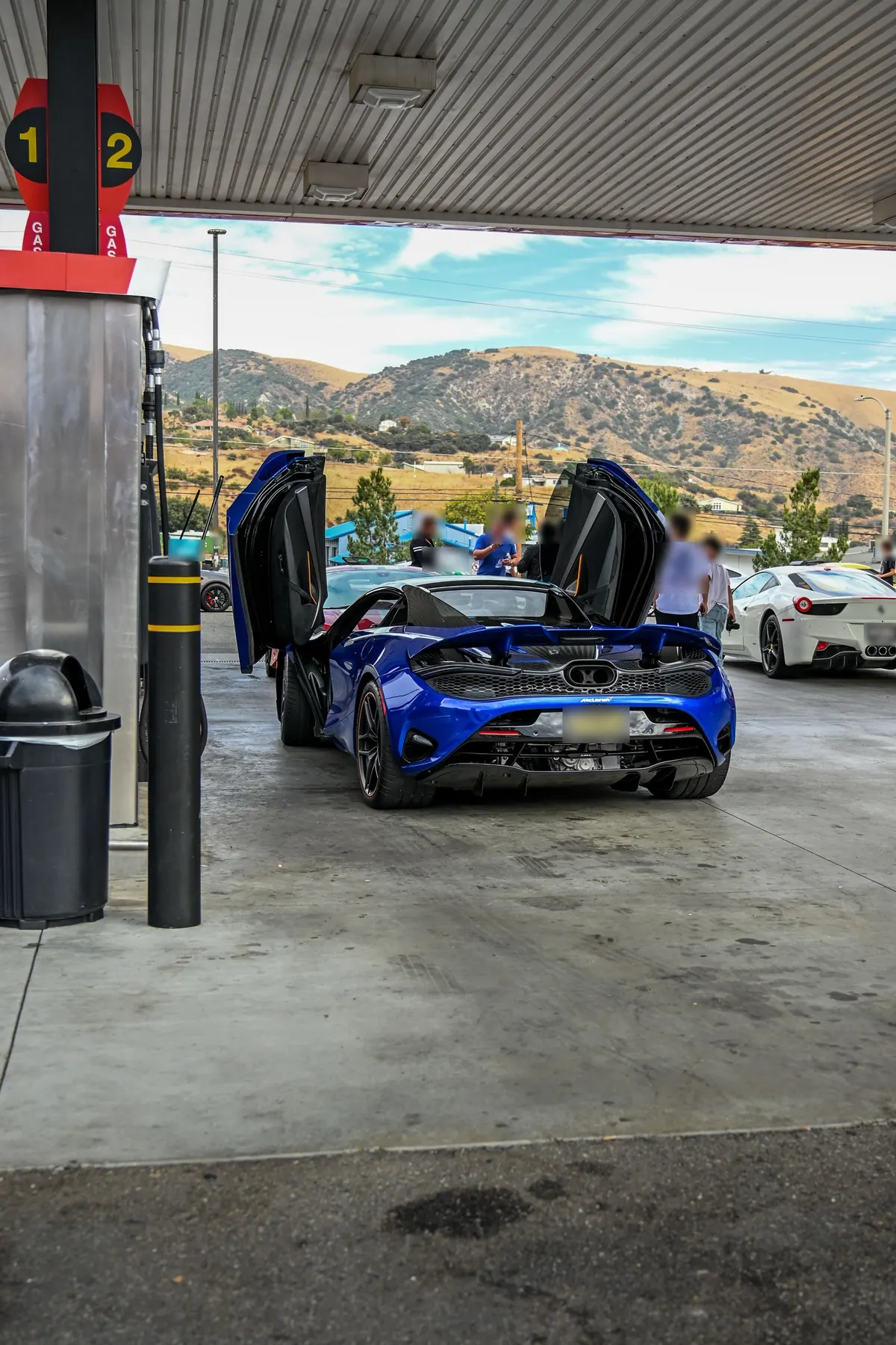 Rear three-quarter view of a blue McLaren 750S with its dihedral doors open at a gas station