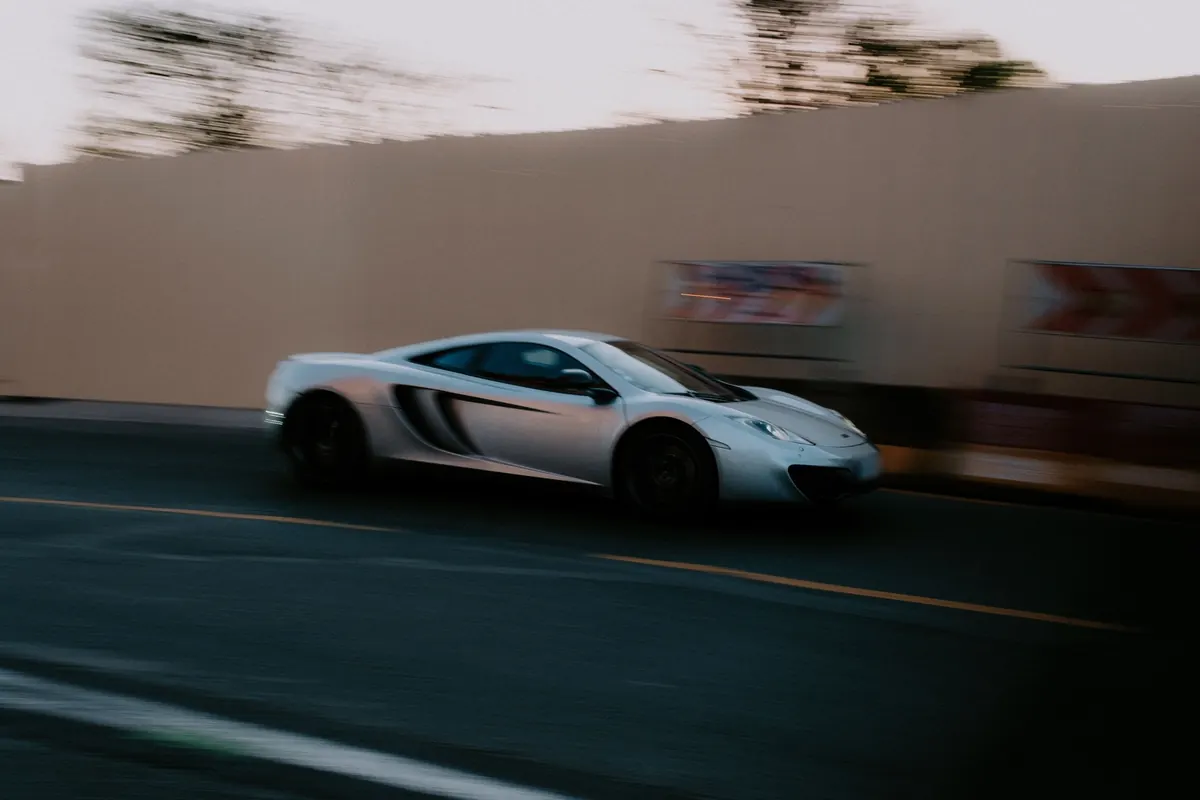 Side view of a silver McLaren MP4-12C driving along a road past a tall fence