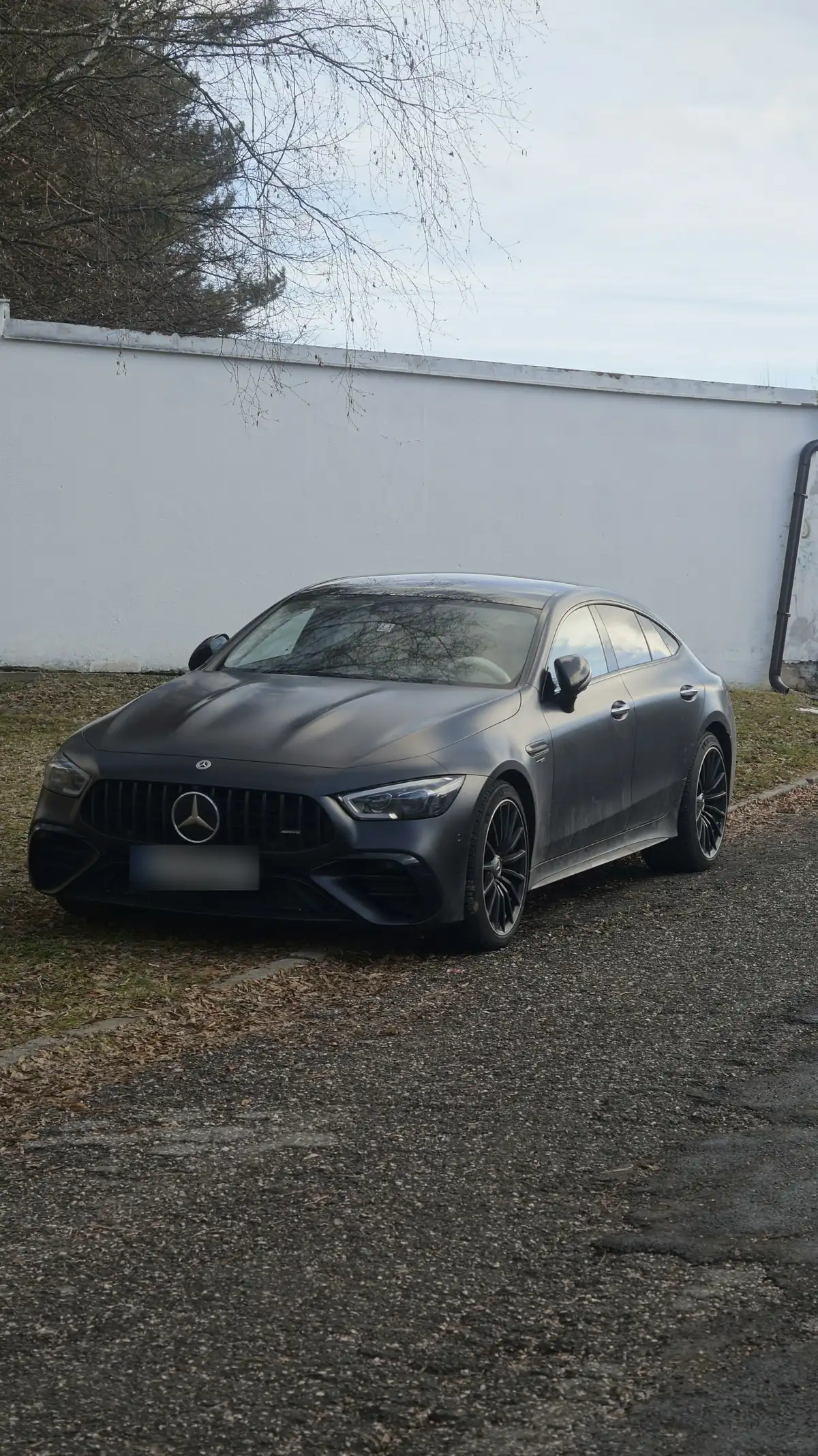 Front three-quarter view of a matte grey Mercedes-AMG GT 53 4-Door Coupé (X290) parked by a white wall