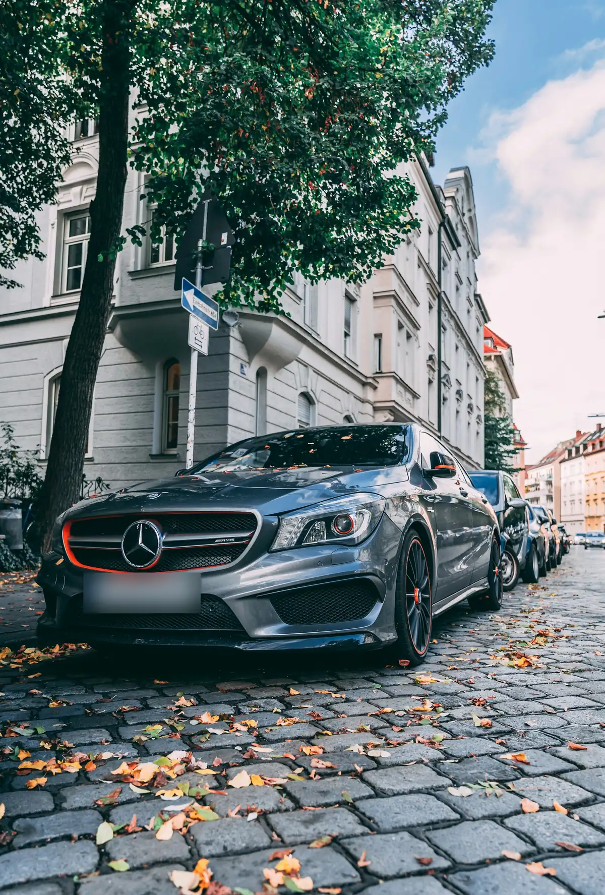Front three-quarter view of a grey Mercedes-AMG CLA45 (C117) with orange accents parked on a cobbled street