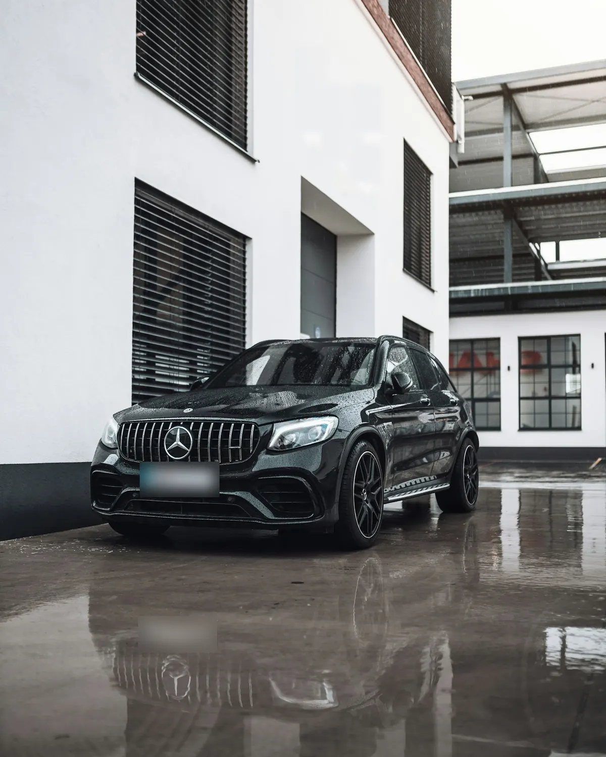 Front three-quarter view of a black Mercedes-AMG GLC63 (X253) on a wet forecourt by a white building