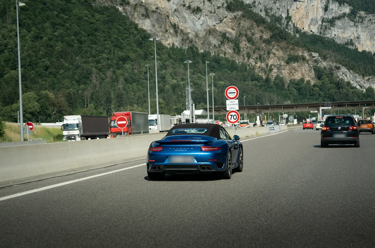 Rear view of a blue Porsche 911 Turbo S (991.1) with its roof up, approaching a highway toll plaza